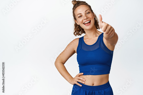 Fototapeta Naklejka Na Ścianę i Meble -  Smiling young spportswoman shows thumbs up. Motivated fitness woman like something, giving compliment, recommending sport gym, white background