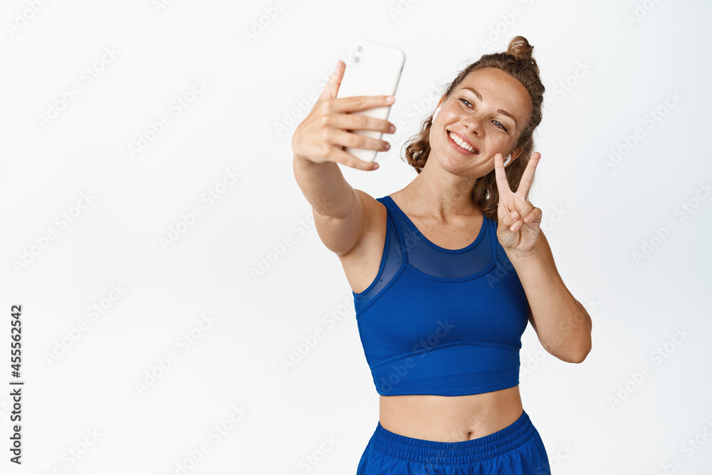 Happy female athlete taking selfie on mobile phone, photographing in headphones and activewear, white background Happy female athlete taking selfie on mobile phone, photographing in headphones and activewear, white background