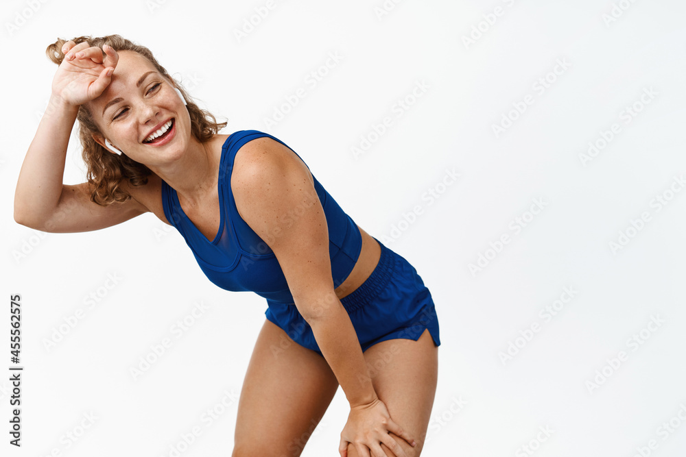 Studio shot of young sportswoman wipe sweat after running, lean on knees and looking behind with satisfied face, reaching goal while jogging, standing against white background Studio shot of young sportswoman wipe sweat after running, lean on knees and looking behind with satisfied face, reaching goal while jogging, standing against white background