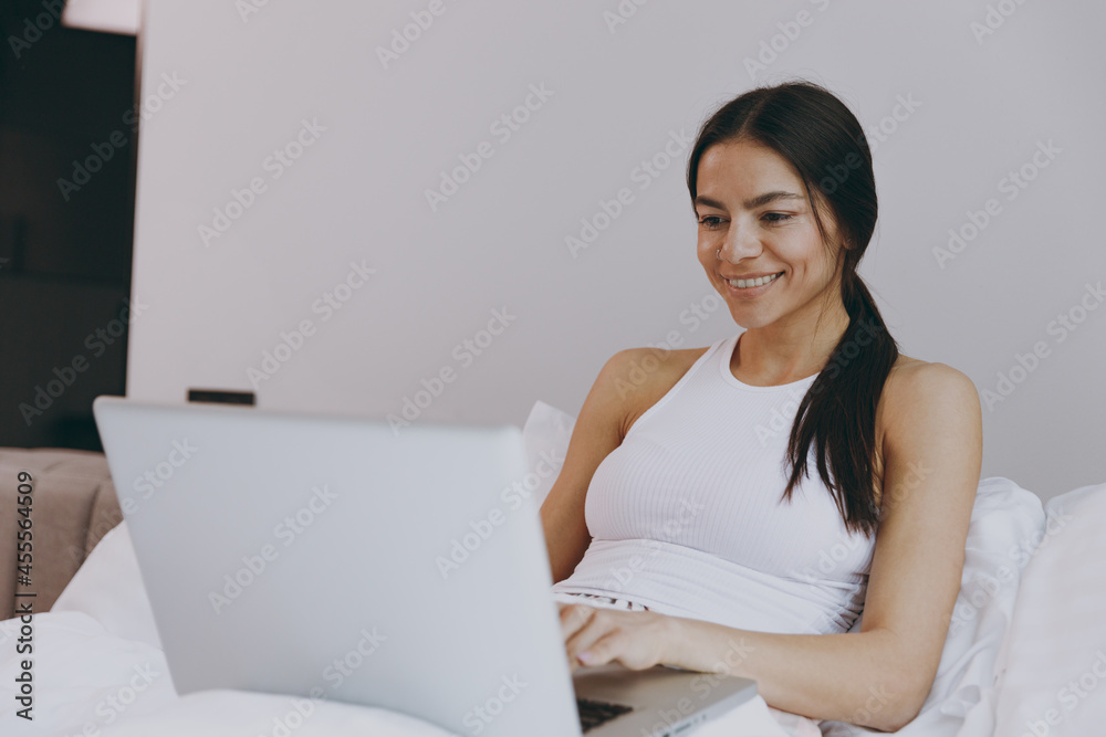 Fascinating charming vivid young woman in white tank top sit in bed use work on laptop pc computer searching browse in bedroom lounge home in own room house wake up dream be lost in reverie good day Fascinating charming vivid young woman in white tank top sit in bed use work on laptop pc computer searching browse in bedroom lounge home in own room house wake up dream be lost in reverie good day