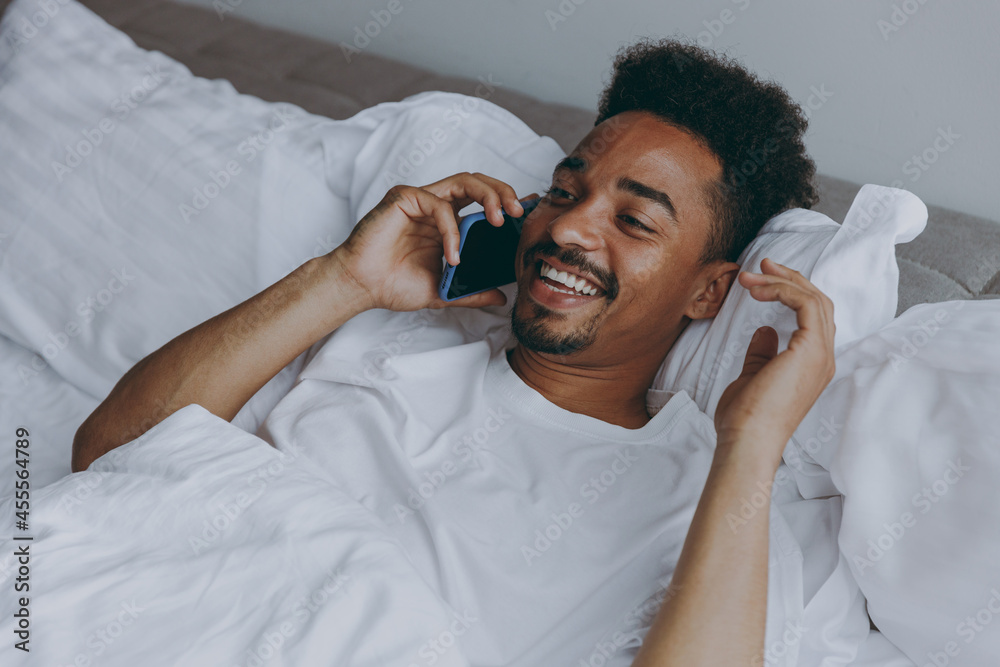 Young satisfied smiling african american man in nightwear talk mobile cell phone lying in bed rest relax spend time in bedroom lounge home in own room house wake up dream be lost in reverie good day. Young satisfied smiling african american man in nightwear talk mobile cell phone lying in bed rest relax spend time in bedroom lounge home in own room house wake up dream be lost in reverie good day.