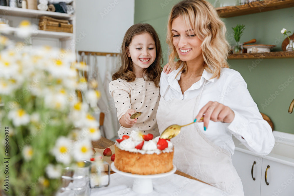 Happy cute chef cook baker mom woman in white shirt work with child baby girl helper decorate pie with cream at kitchen table home. Cooking food concept Mommy little kid daughter prepare fruit cake