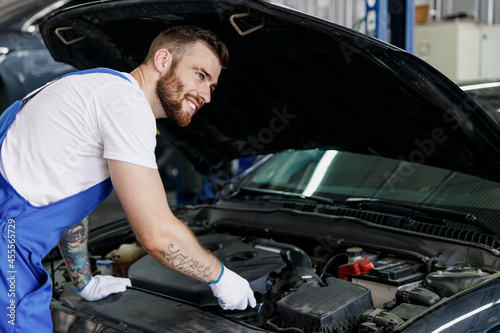 Minded troubleshooter young male professional technician car mechanic man in denim blue overalls white t-shirt fixing problem with raised hood work in light modern vehicle repair shop workshop indoor.