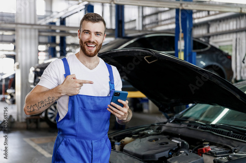 Young professional technician car mechanic man in blue overalls white t-shirt use hold point finger on mobile cell phone fix problem with raised hood bonnet work in vehicle repair shop workshop inside