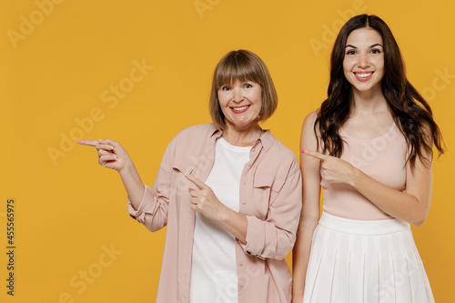 Two young smiling fun happy daughter mother together couple women wearing casual beige clothes pointing on workspace area copy space mock up isolated on plain yellow color background studio portrait photo