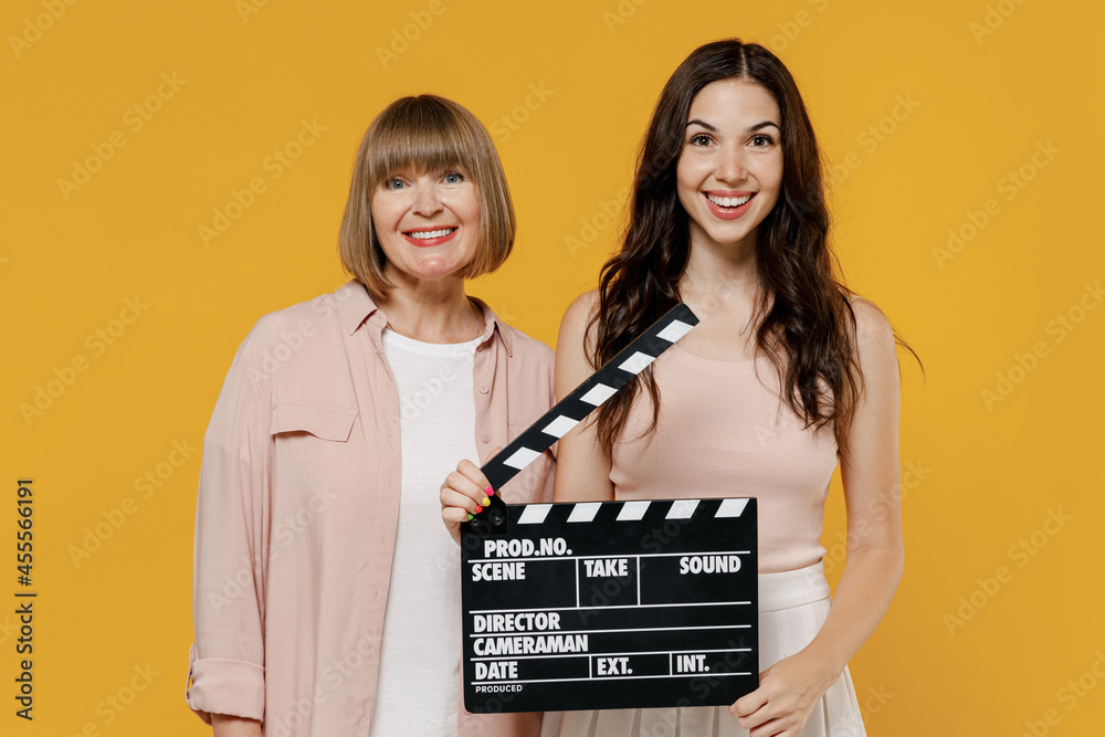 Two young smiling happy daughter mother together couple women wearing casual beige clothes holding classic black film making clapperboard isolated on plain yellow color background studio portrait