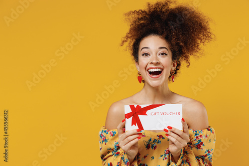 Young happy smiling fun woman 20s with culry hair in casual clothes hold gift certificate coupon voucher card for store isolated on plain yellow background studio portrait. People lifestyle concept