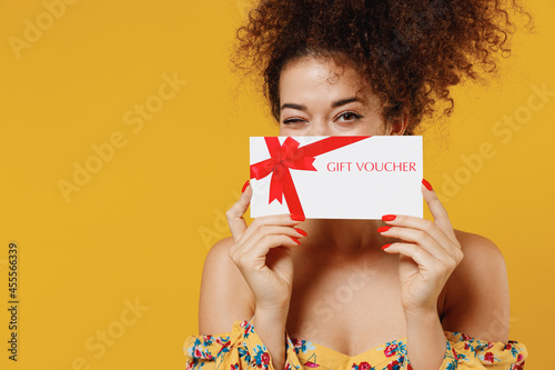 Young happy smiling fun cheerful woman 20s with culry hair in casual clothes hold cover face with gift certificate coupon voucher card for store isolated on plain yellow background studio portrait