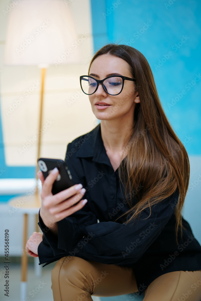 Stunning beautiful girl smiling and poses to the camera in modern glasses. Woman sitting and looking at the smartphone. Light interior colors. Closeup