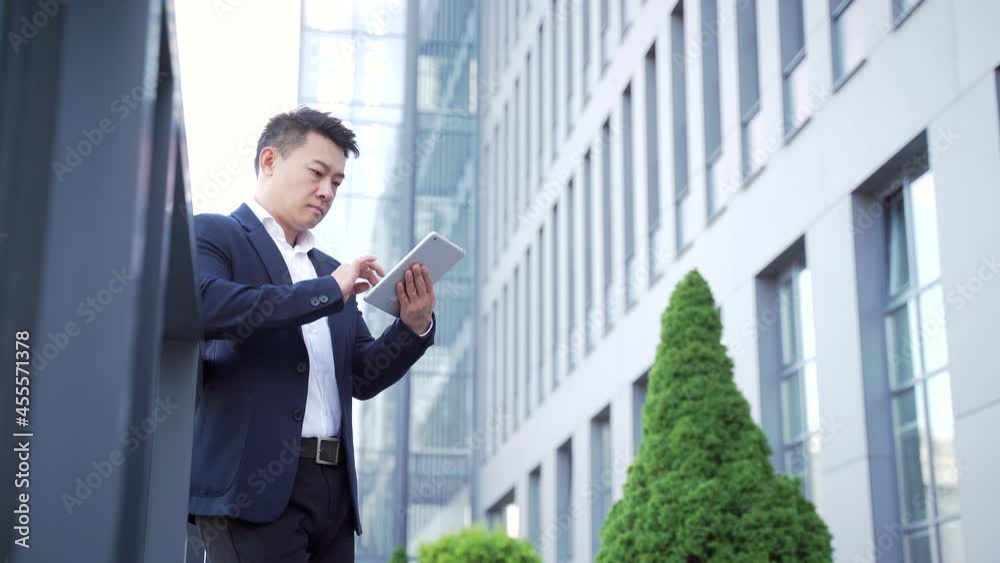 young asian businessman standing near the railing of terrace a modern office building using a digital tablet in his hands. Successful business man on urban background on city street. outside. Outdoors young asian businessman standing near the railing of terrace a modern office building using a digital tablet in his hands. Successful business man on urban background on city street. outside. Outdoors