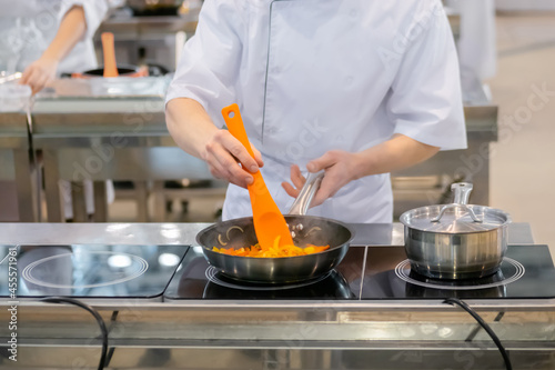 Chef hands holding silicone spatula and cooking vegetables - sliced bell pepper and onion in frying pan with oil on electric stove - front view. Professional cooking  gastronomy and food concept