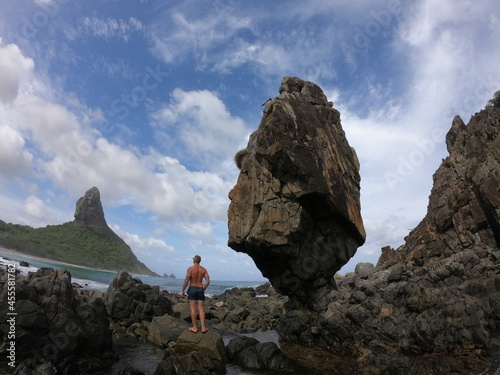 Man in swim suit standing beside a huge rock at the rocky beaches of Fernando de Noronha, Pernambuco, Brazil photo