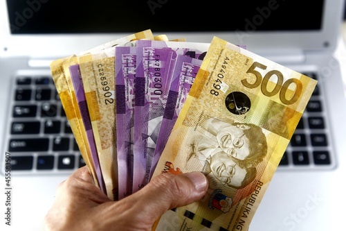 Person holding 100 and 500 Philippine peso banknotes beside laptop photo