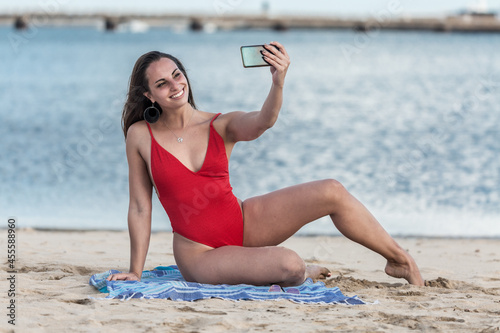 Woman in red swimsuit sitting on blue mat on beach taking selfie