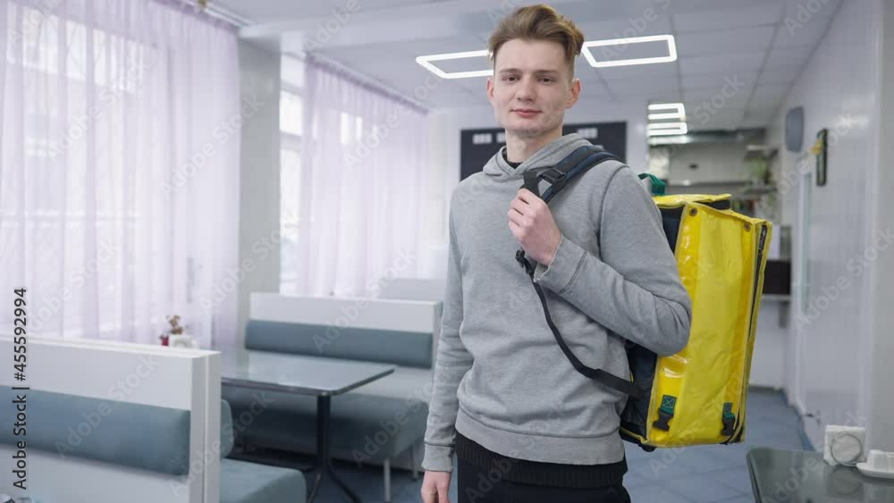 Confident young male courier hanging food delivery bag on shoulder looking at camera smiling. Portrait of Caucasian man posing in cafeteria indoors. Delivering and employment concept Confident young male courier hanging food delivery bag on shoulder looking at camera smiling. Portrait of Caucasian man posing in cafeteria indoors. Delivering and employment concept