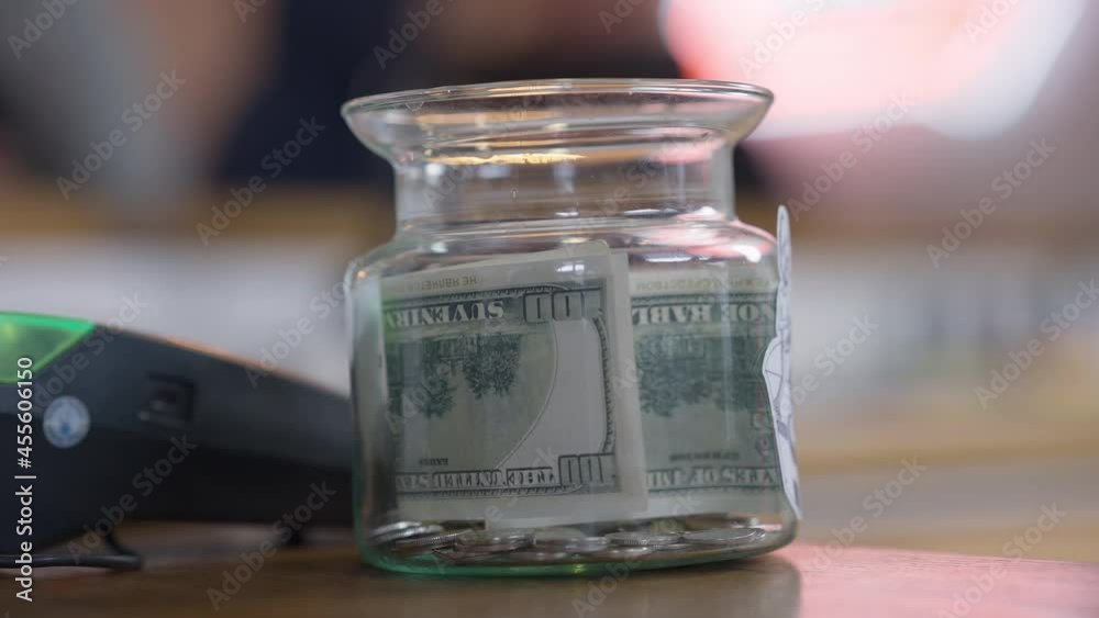 Close-up tips jar with female Caucasian hand putting cash inside. Unrecognizable cafe visitor leaving money for tips in cafeteria indoors Close-up tips jar with female Caucasian hand putting cash inside. Unrecognizable cafe visitor leaving money for tips in cafeteria indoors