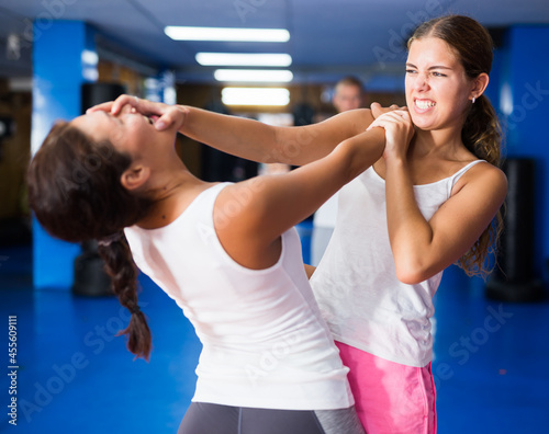 Concentrated young girl practicing effective self defence techniques while sparring in training room, blocking hand of female opponent and launching palm blow in face