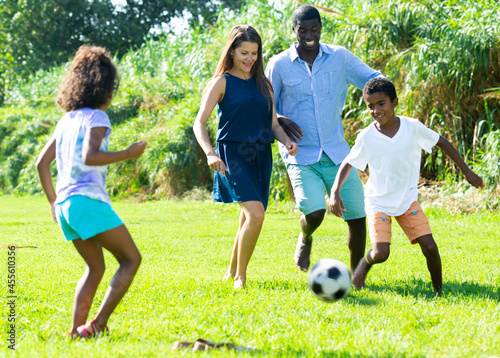 Happy friendly interracial family with preteen children playing ball on green lawn in summer city park