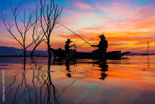 Asian fisherman on wooden boat casting a net for catching freshwater fish in nature river in the early morning before sunrise.