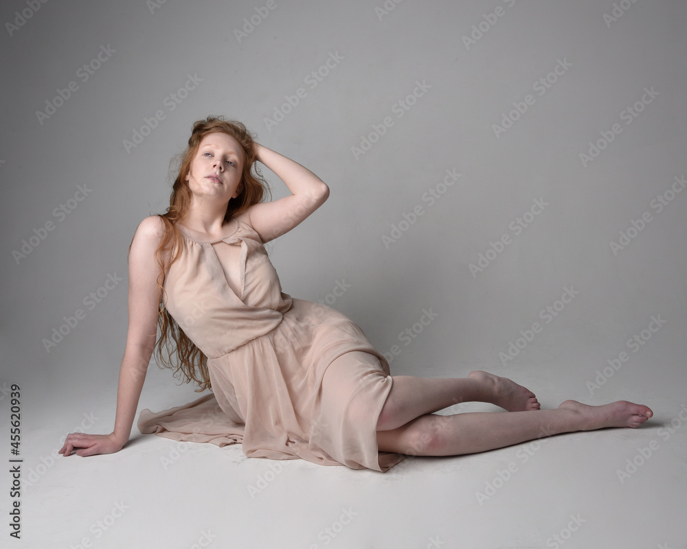 Naklejka premium Full length portrait of pretty red haired woman dancer, wearing skin toned flowing fairy dress. Sitting gestural poses isolated on studio background.