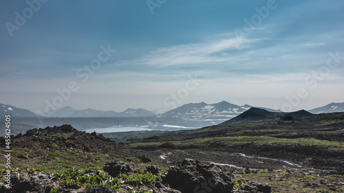 Scant vegetation and volcanic rocks are visible on the mountain slope. In the distance, you can see a lake, a picturesque mountain range against the blue sky. Kamchatka photo