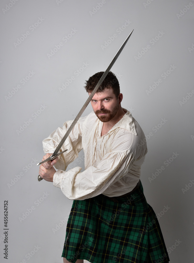 Close up portrait of handsome brunette man wearing Scottish kilt and renaissance white pirate blouse shirt. Holding a sword weapon, action pose isolated against studio background. Close up portrait of handsome brunette man wearing Scottish kilt and renaissance white pirate blouse shirt. Holding a sword weapon, action pose isolated against studio background.