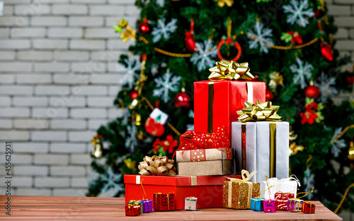 Colorful small and big present gift boxes with shiny ribbon bow tie placed on corner of wooden table in front fully decorative beautiful Christmas eve pine tree and brick wall in blurred background