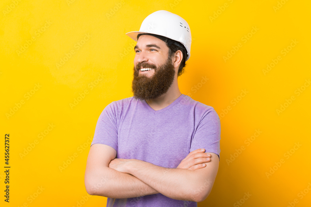 Confident bearded man wearing hard hat is standing with crossed arms over yellow background.
