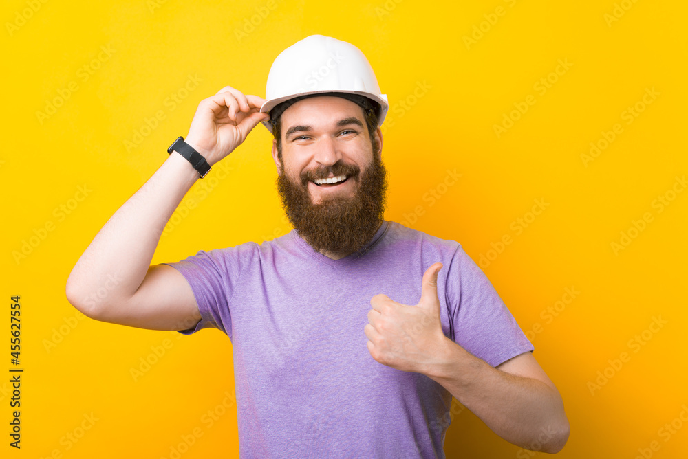 Wearing protection hard hat is a good idea - says bearded man who has one and shows thumb up over yellow background.