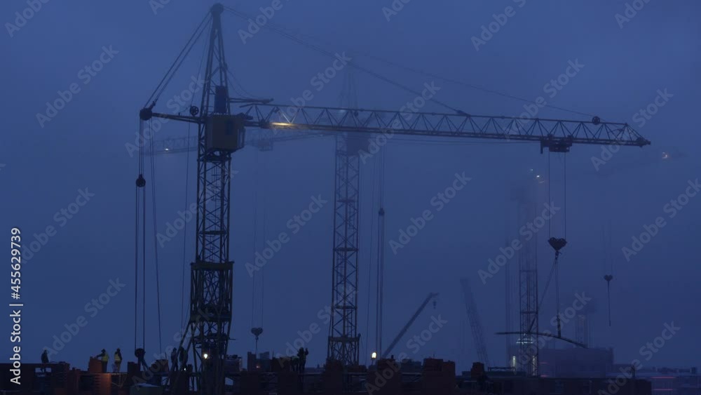 Construction site at dusk with purple sky. Silhouette of high tower cranes works on high-rise residential building site, lifts load. Construction site at dusk with purple sky. Silhouette of high tower cranes works on high-rise residential building site, lifts load.
