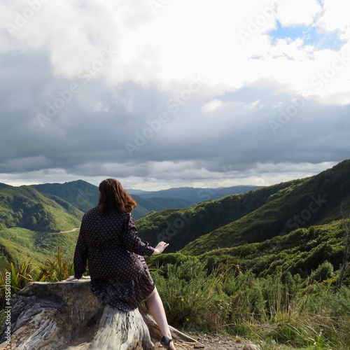 A view over the Rimutaka mountain range in New Zealand on a dark, cloudy day. A woman sits atop a log in the foreground, looking at the view.