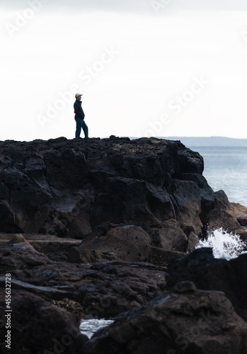 Man standing on rocks looking at the sea. Waves crashing onto the rocks  Takapuna and Milford walkway. Vertical format.