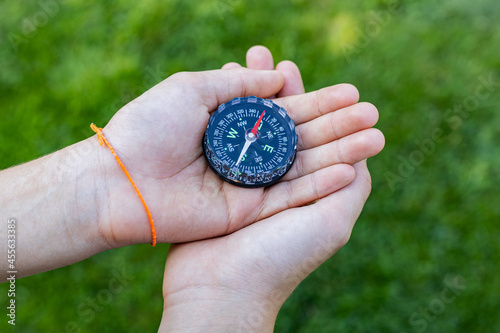 Girl holding the compass. Hands of a teenager girl holding a liquid compass. Green grass background. Copy space. Red compass needle points north. Orienteering on the ground. Selecting the direction