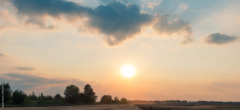 A wide-angle landscape of summer evening farm field, under the magnificent sunset sky with clouds. Harvesting season.