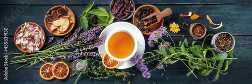 Tea panorama. Herbs, flowers and fruit around a cup of tea, an overhead flat lay shot on a dark rustic wooden background