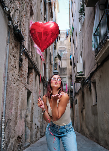 Woman blowing an air kiss to the camera while holding a red heart shape balloon outdoors.