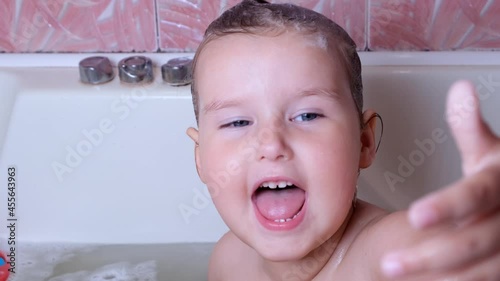 Little funny girl 3 years old bathes and washes with foam on her head in water in the bathroom at home. Baby body care concept, hygiene.