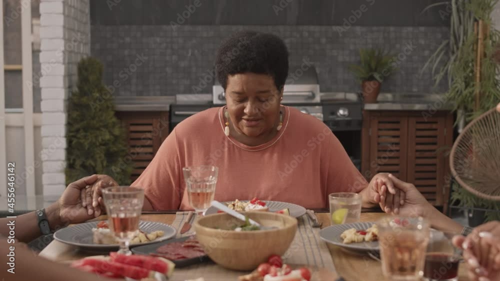 Chest-up of mature African-American woman sitting at table, holding hands together with cropped family members, praying before having dinner Chest-up of mature African-American woman sitting at table, holding hands together with cropped family members, praying before having dinner
