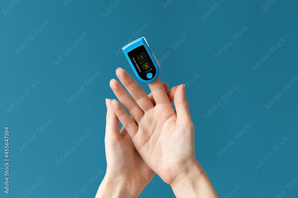 Close up of females hand with a pulse oximeter placed on the index finger. Blue background. The concept of measuring oxygen in the blood Close up of females hand with a pulse oximeter placed on the index finger. Blue background. The concept of measuring oxygen in the blood