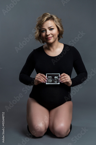 Beautiful pregnant woman in black bodysuit with ultrasound photo. Smiling blonde. Happiness is waiting for the baby. Gray background. Vertical.