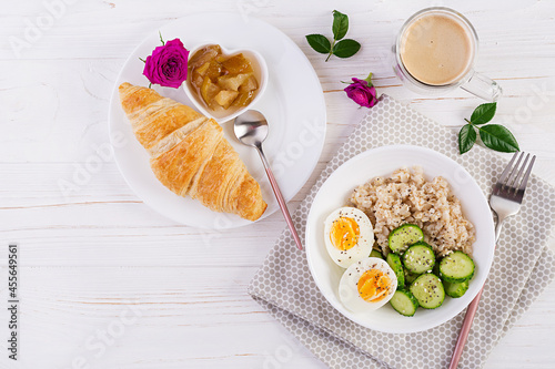 Breakfast oatmeal porridge with boiled egg, cucumber and croissant, jam, coffee. Healthy balanced food. Top view, flat lay
