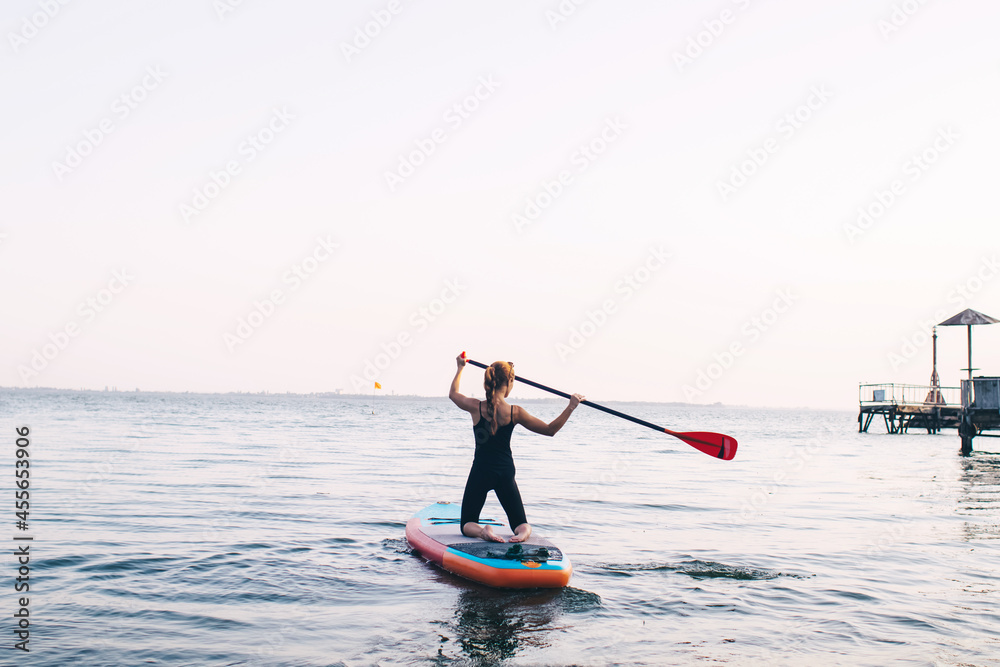 Naklejka premium Close-up of a girl floating on a sup board. The concept of water sports, relaxation and self-immersion. Lonely woman alone on a board against a background of water and a pier.