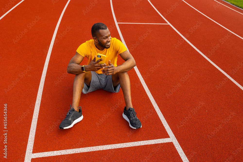 Happy attractive male athlete sitting at the stadium track, relaxing, taking a break after running. Handsome african american man using smartphone, browsing internet, smiling, workout outdoors concept