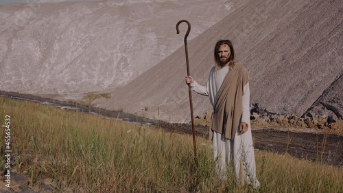 Jesus Christ with a staff in robe stands on the mountain and looks into the distance. There are bruises on his face, wounds on his hands.