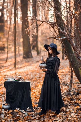 A witch in the autumn forest. A woman in a black long dress cooking a potion is preparing for Halloween.