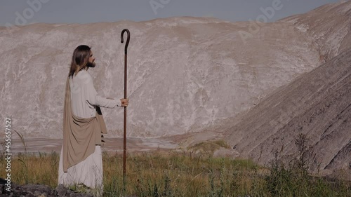 Jesus Christ with a staff in robe stands on the mountain and looks into the distance. There are bruises on his face, wounds on his hands.