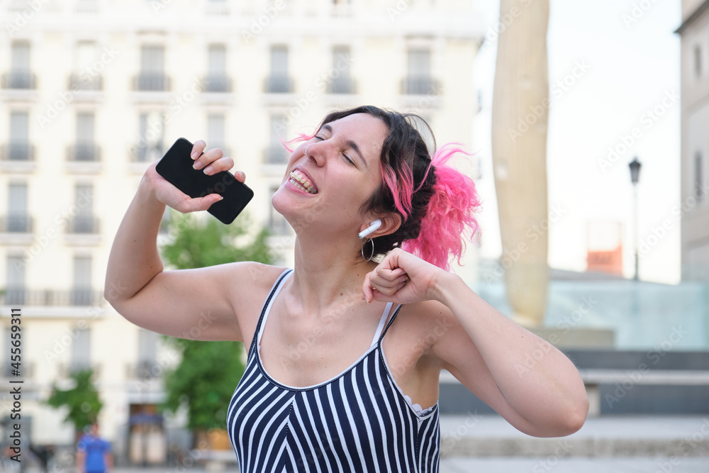 Young caucasian woman with pink hair dancing in street with earbuds and phone.
