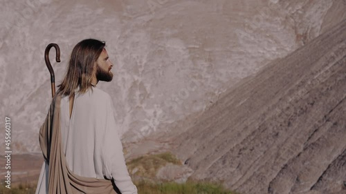 Jesus Christ with a staff in robe stands on the mountain and looks into the distance. There are bruises on his face, wounds on his hands.