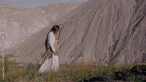 Jesus with a staff walks on the grass against the background of the year. The wind is blowing, the hair is developing. High quality 4k footage