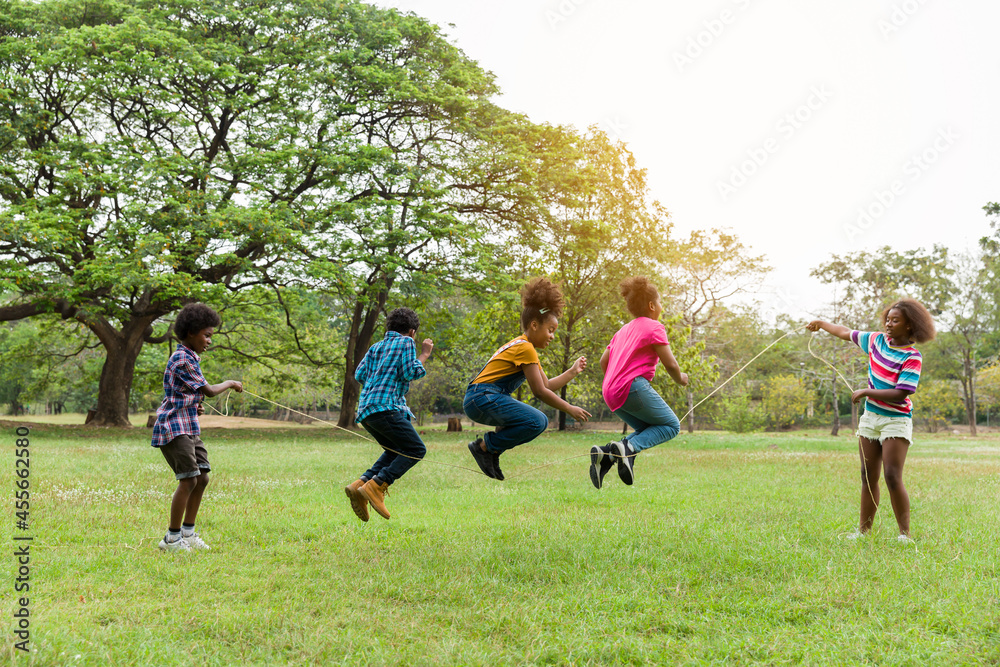 Group of African American children having fun jumping over the rope in the park. Cheerful kid jumping over the rope outdoor. Happy black people enjoying playing together on green grass - obrazy, fototapety, plakaty 